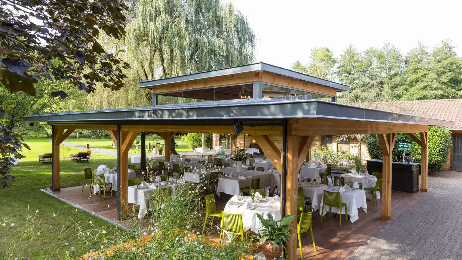 A covered wooden terrace with tables set for a meal, surrounded by trees and greenery.