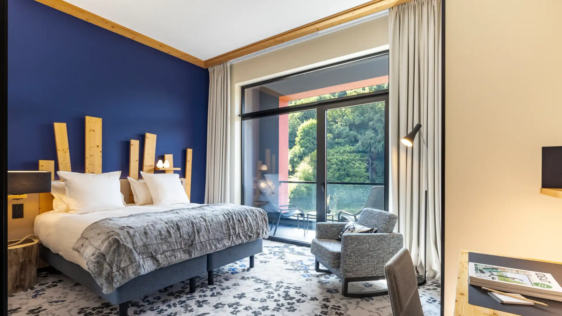 Room with a double bed, a bedside table, an armchair, and a large window opening onto a terrace with a view of trees.