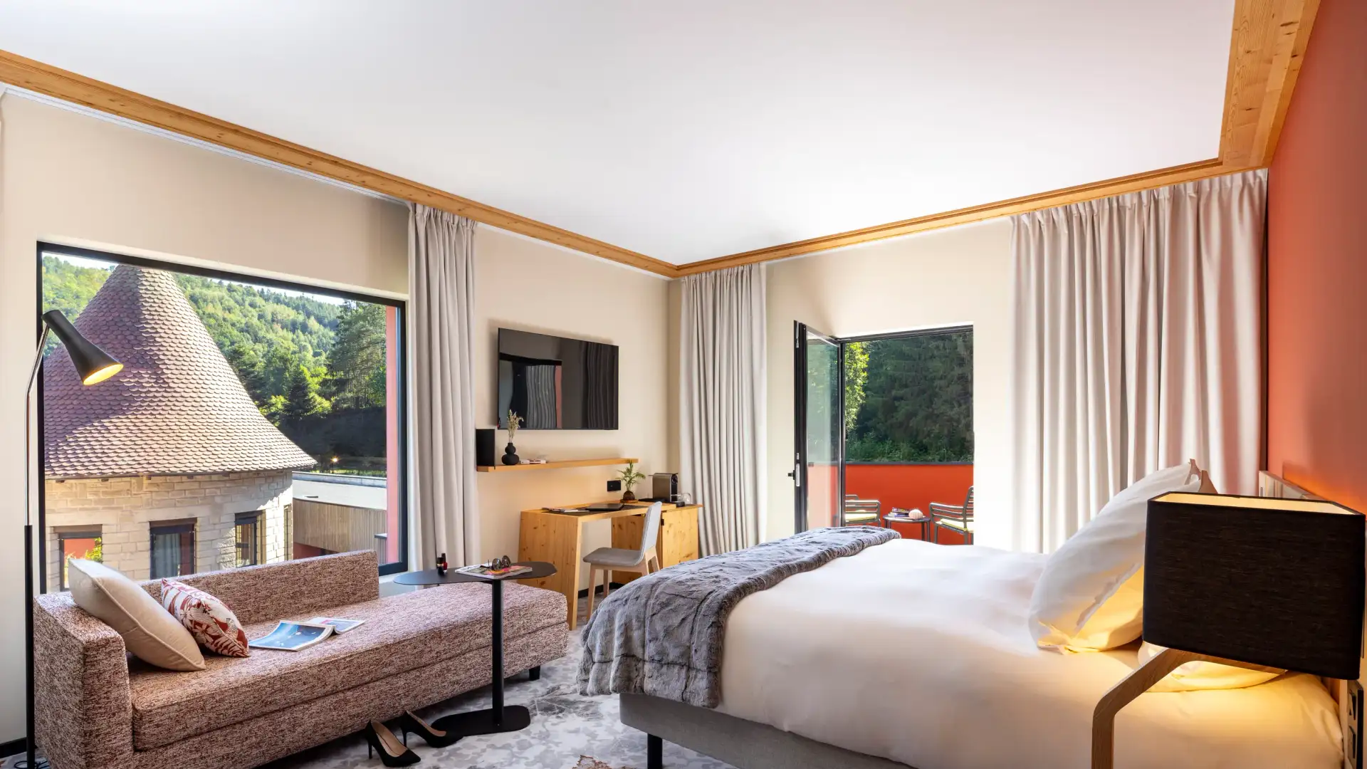 Helles Zimmer mit Bett, Sofa, Schreibtisch und Blick auf traditionelle Dächer und einen Wald