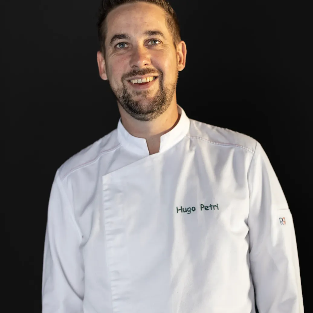 A man in a chef's jacket with the name "Hugo Petri" on his chest smiles at the camera against a black background.