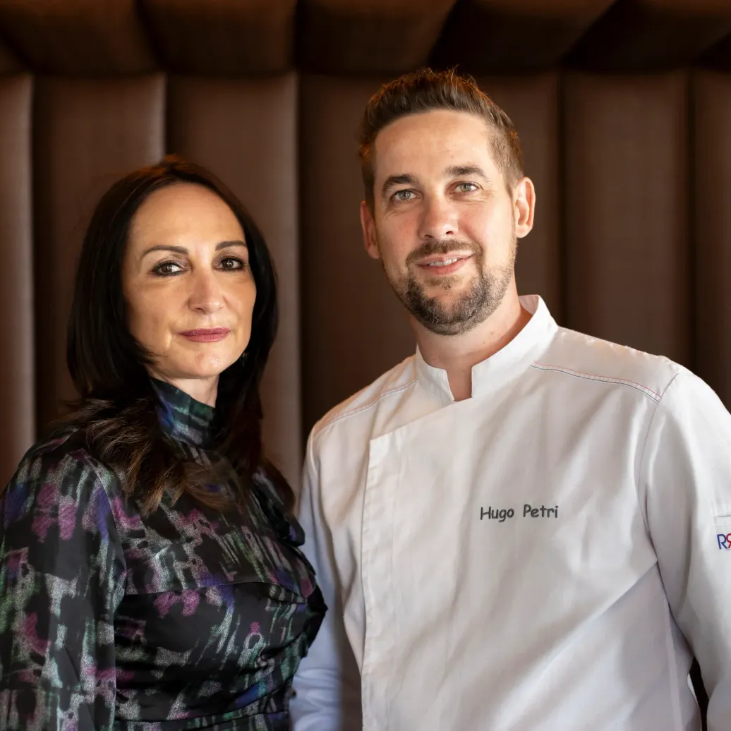 Two people standing side by side, including a man in a chef's jacket with an embroidered name, in front of a dark wooden background.