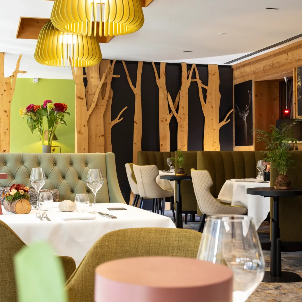 Elegant restaurant with set tables, green banquettes, and golden pendant lights, wooden walls, and decorative plants.