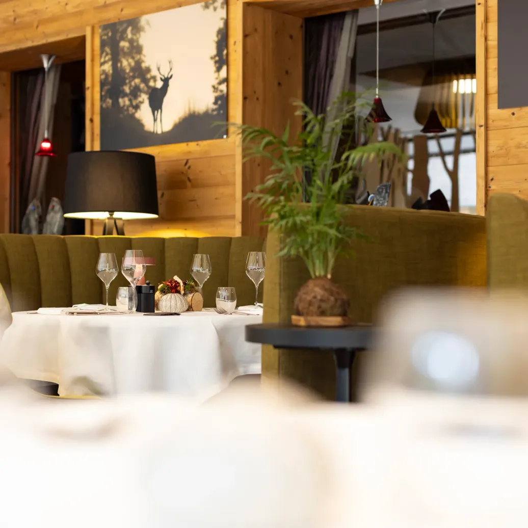 Table set with glasses and white tablecloth in a wooden interior setting with green seats and plants.