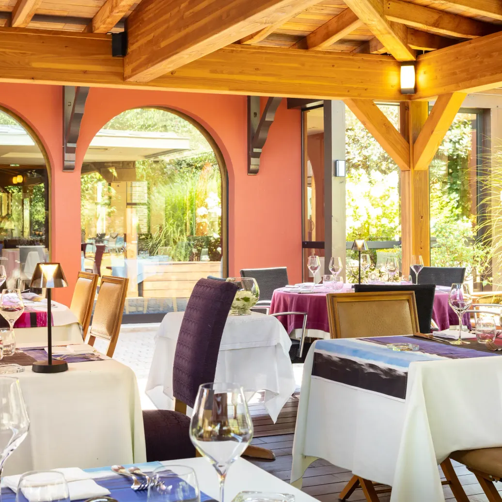 Restaurant terrace with set tables, white tablecloths, and wine glasses, under a wooden structure with orange arches.