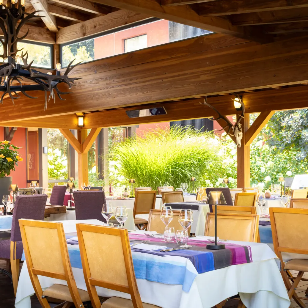 Outdoor dining area with set tables, wooden chairs, and floral decorations, under a wooden pergola with suspended deer antlers.