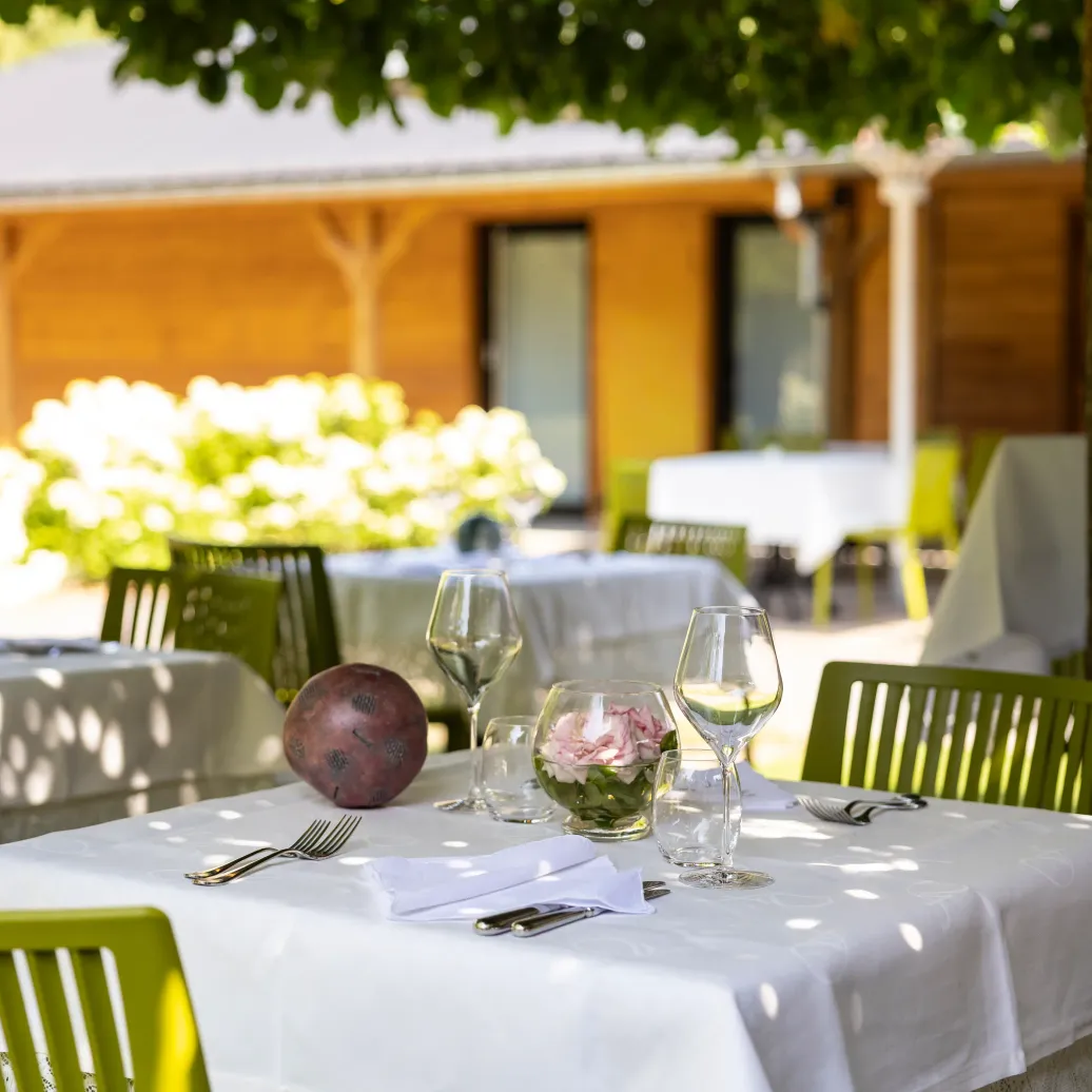 Table set outdoors with white tablecloth, wine glasses, flower vase, and cutlery, under trees and next to other similar tables.