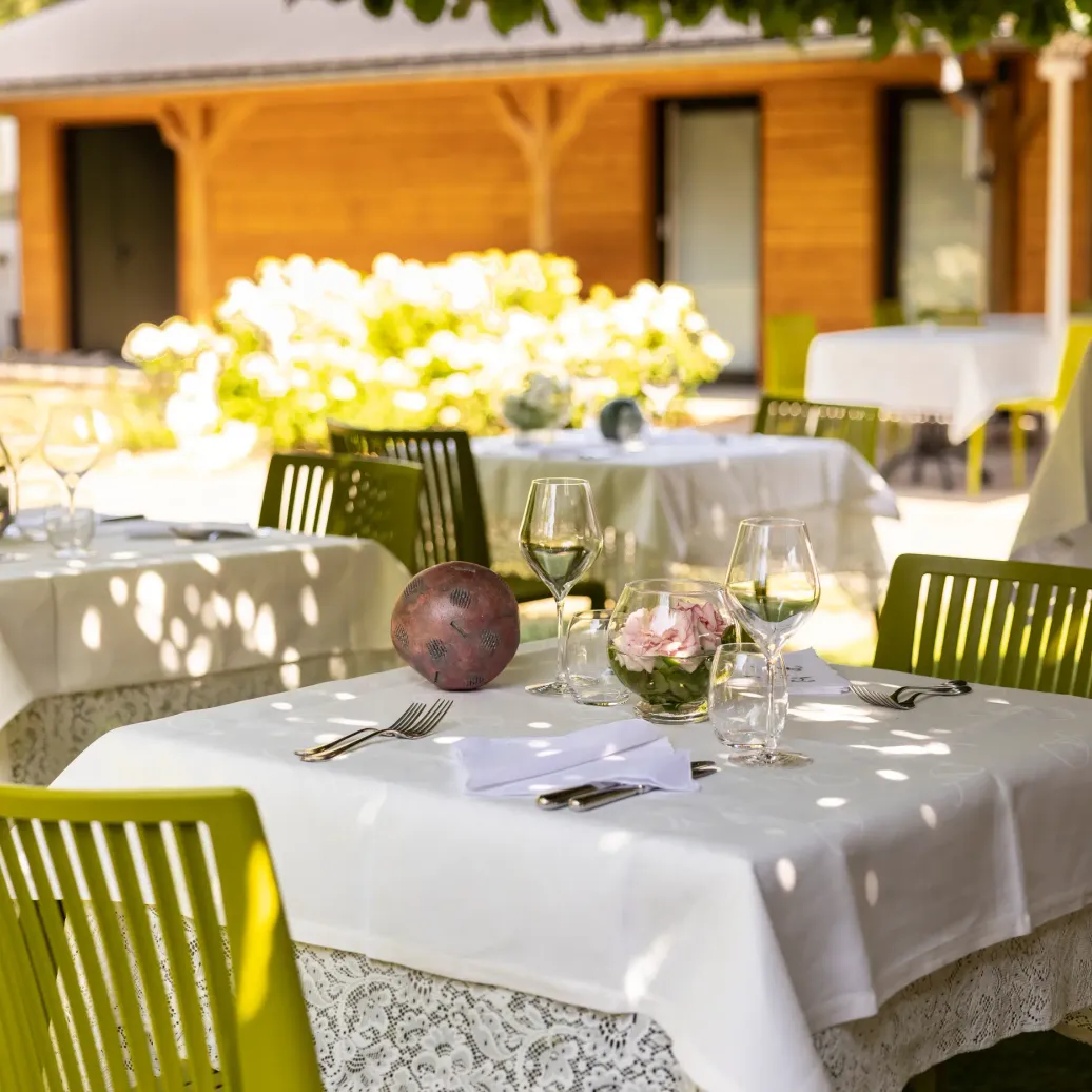 Outdoor tables with white tablecloths, wine glasses, cutlery, and floral decorations under a wooden awning.