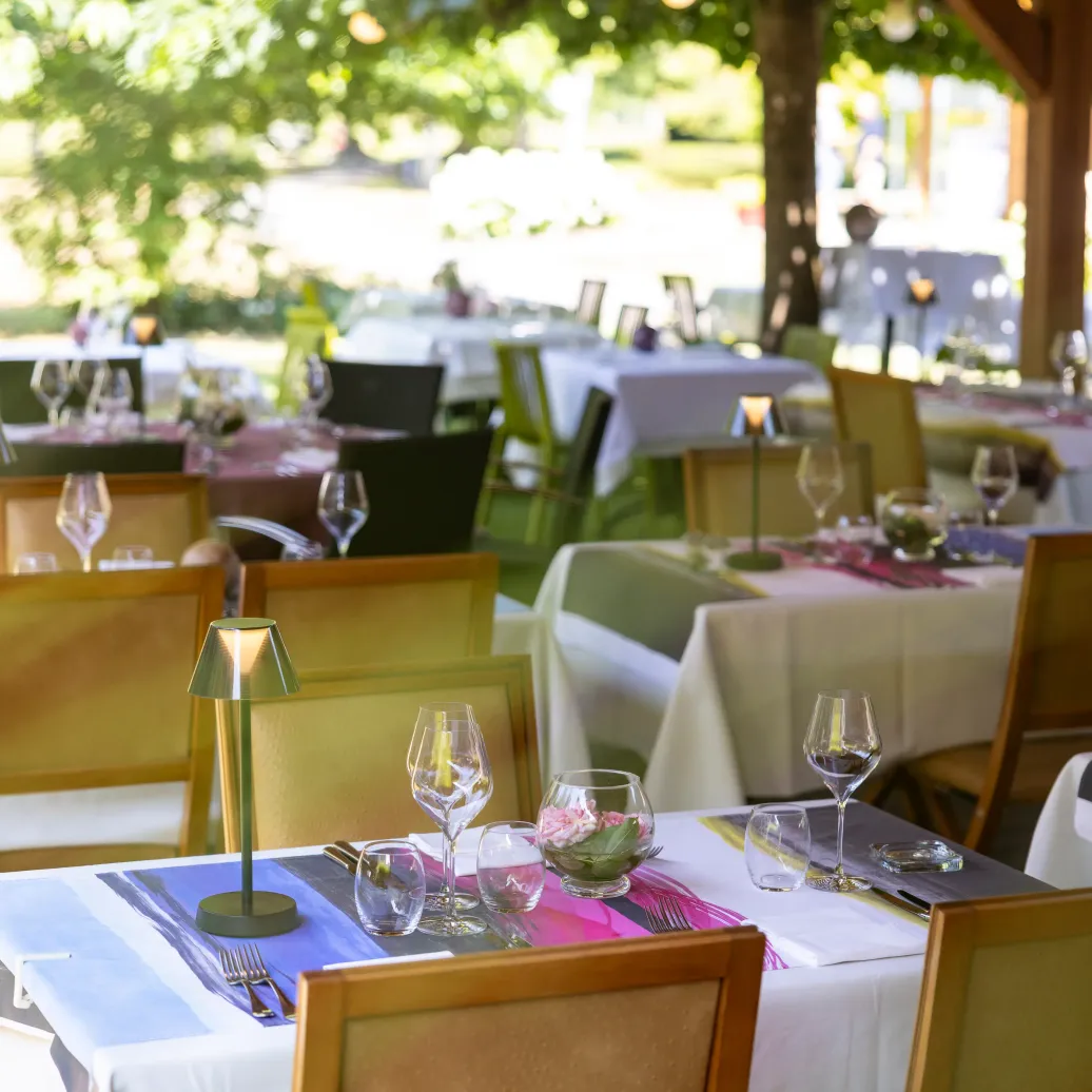 A restaurant terrace with set tables, wine glasses, and candles, surrounded by trees and greenery.