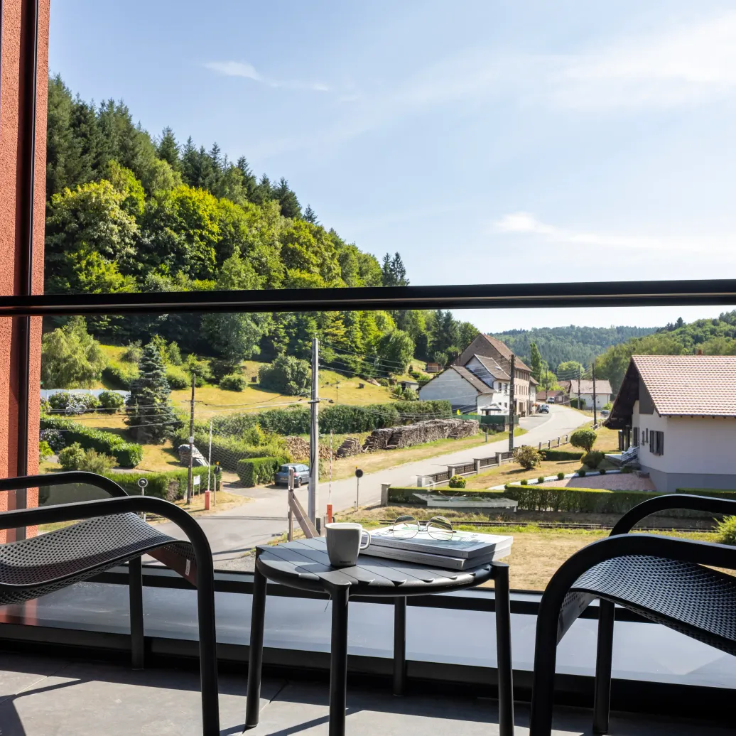 View from a balcony with a table, chairs, and a cup, overlooking a village with houses and greenery in the background.