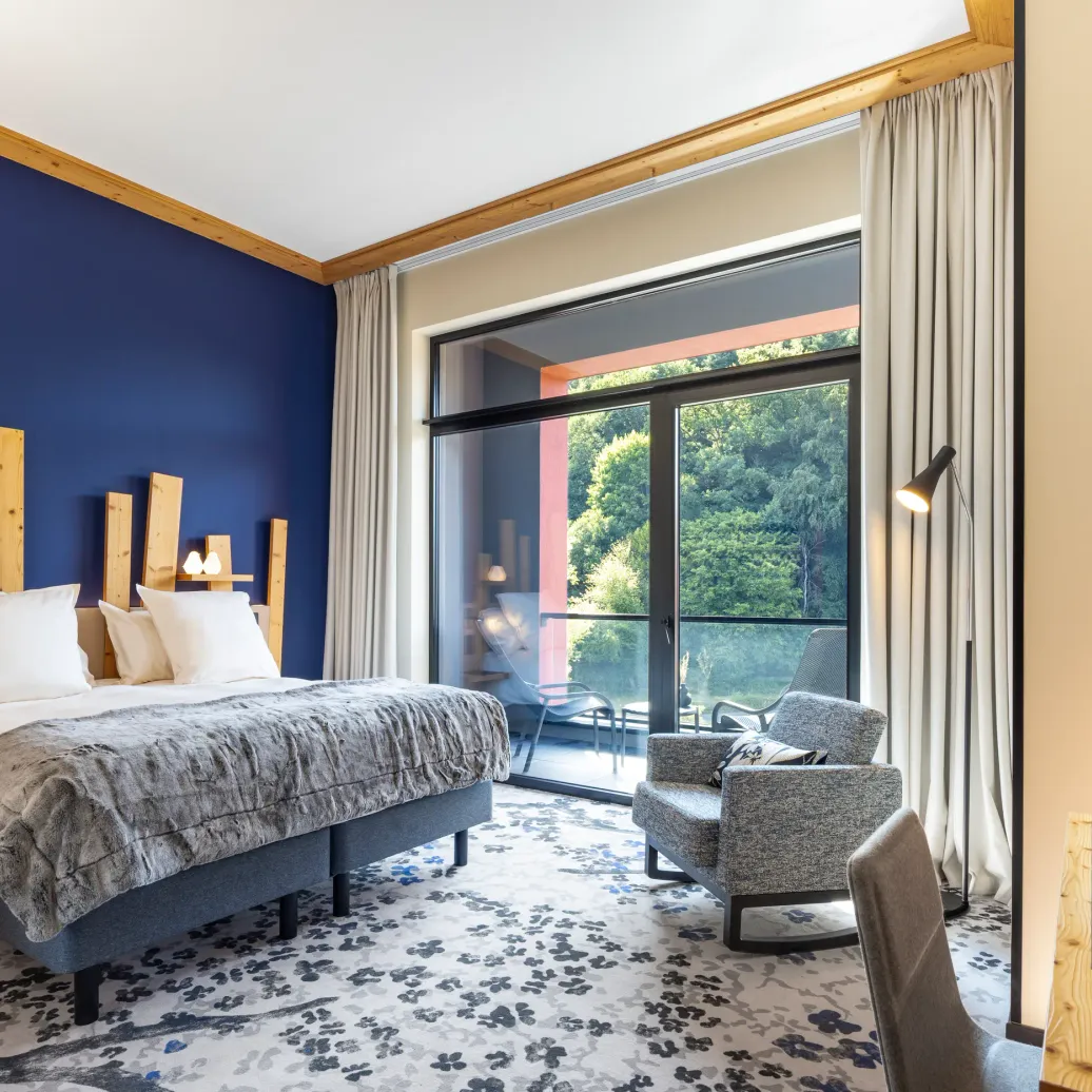 Room with a double bed, a bedside table, an armchair, and a large window opening onto a terrace with a view of trees.