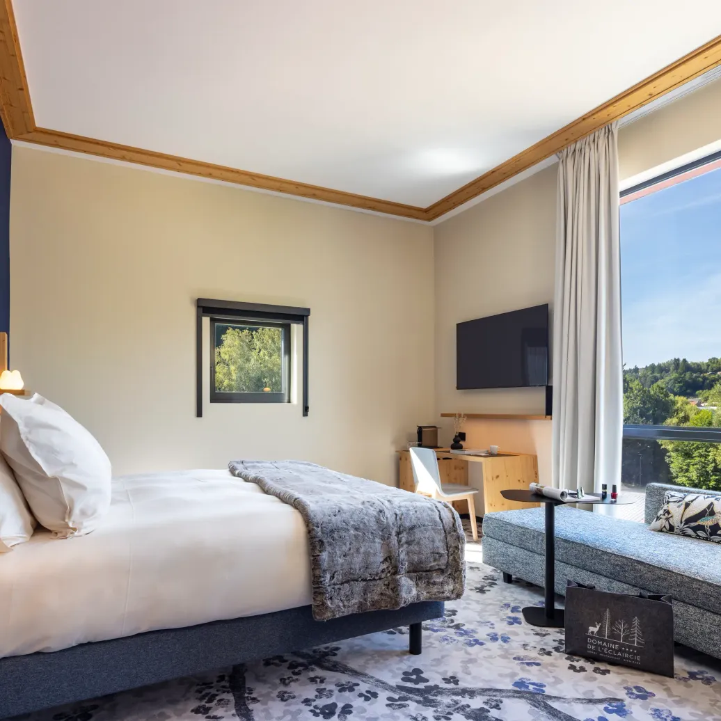 Room with a bed, a bedside table, a sofa, and a large window offering a view of a forest.