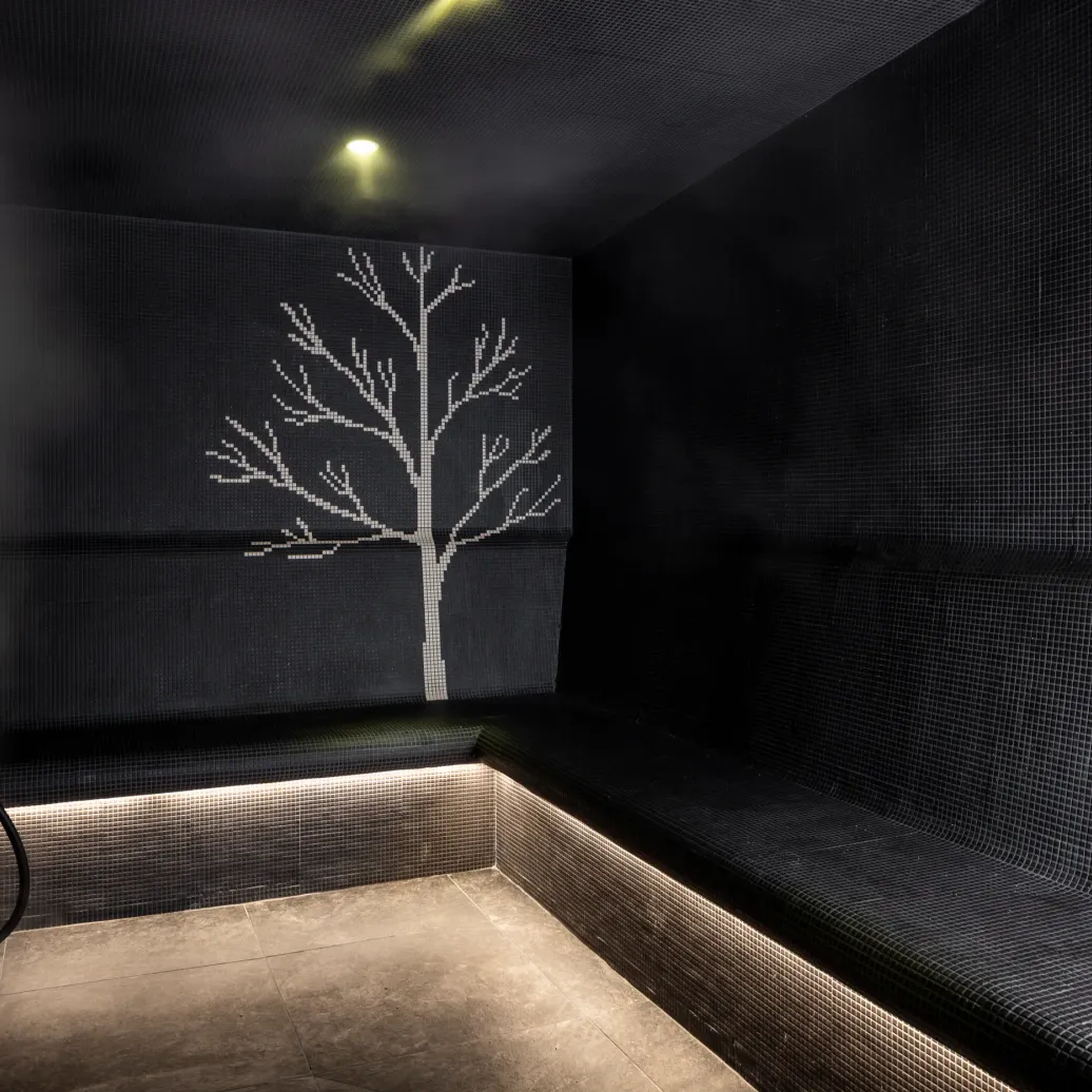 Black shower wall with a mosaic of small tiles depicting a stylized white tree, lit by soft ceiling light