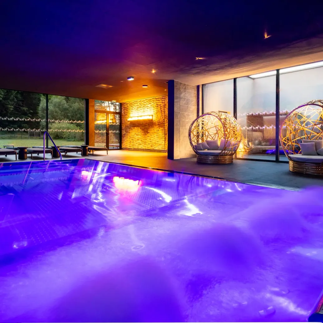 Indoor pool lit in purple with sun loungers and spherical wicker armchairs illuminated nearby, surrounded by large glass windows overlooking a garden