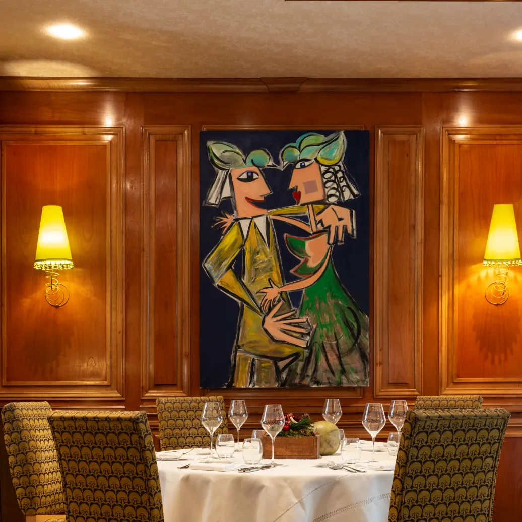 Table set with glasses and cutlery, surrounded by patterned chairs, in a dining room with a wall painting depicting a couple and colorful stained-glass windows on the ceiling.