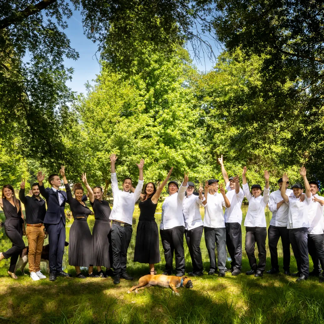 A group of people raises their arms in the air in a tree-lined park near a body of water.