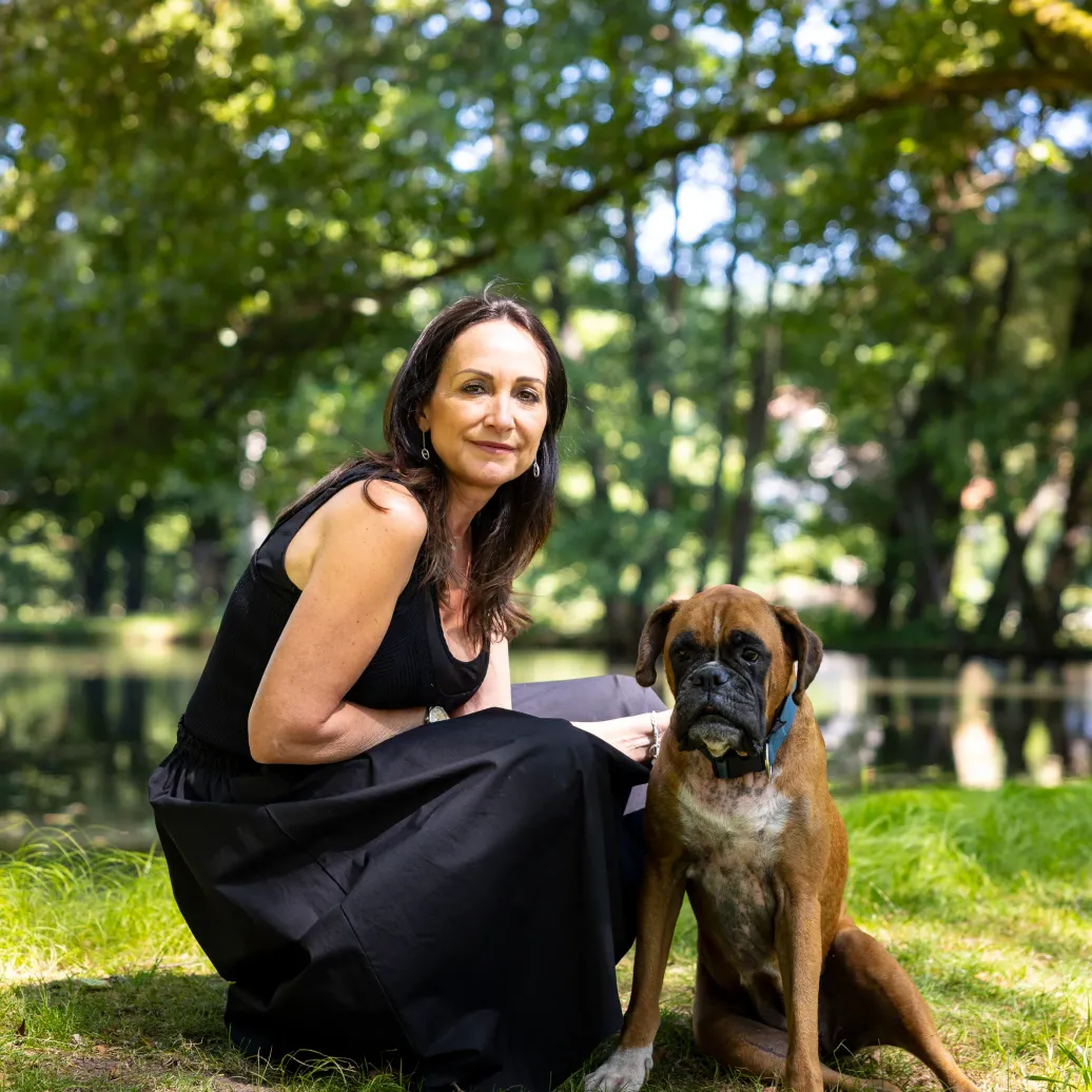Woman in a black dress sitting in the grass with a boxer dog next to her in a tree-lined park