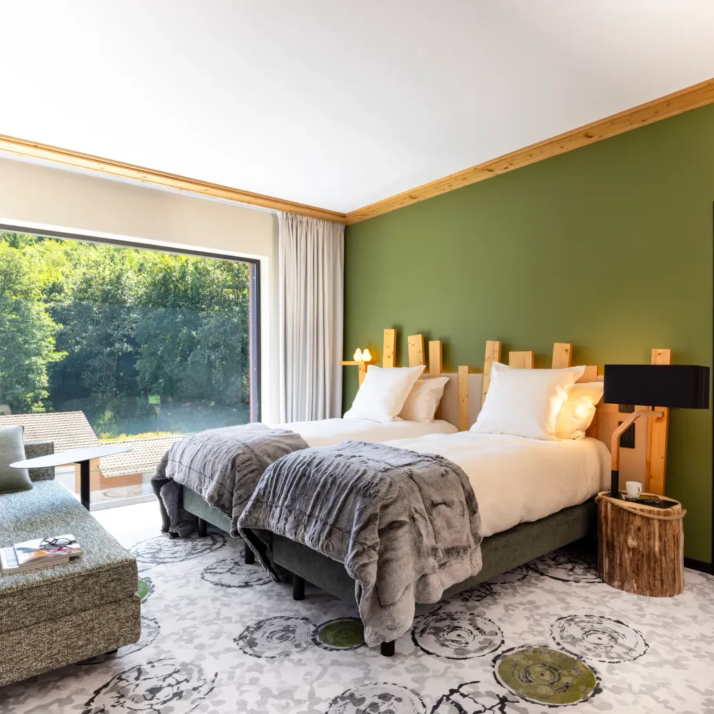 Room with two beds, a sun lounger, a wooden bedside table and a large window overlooking a forest