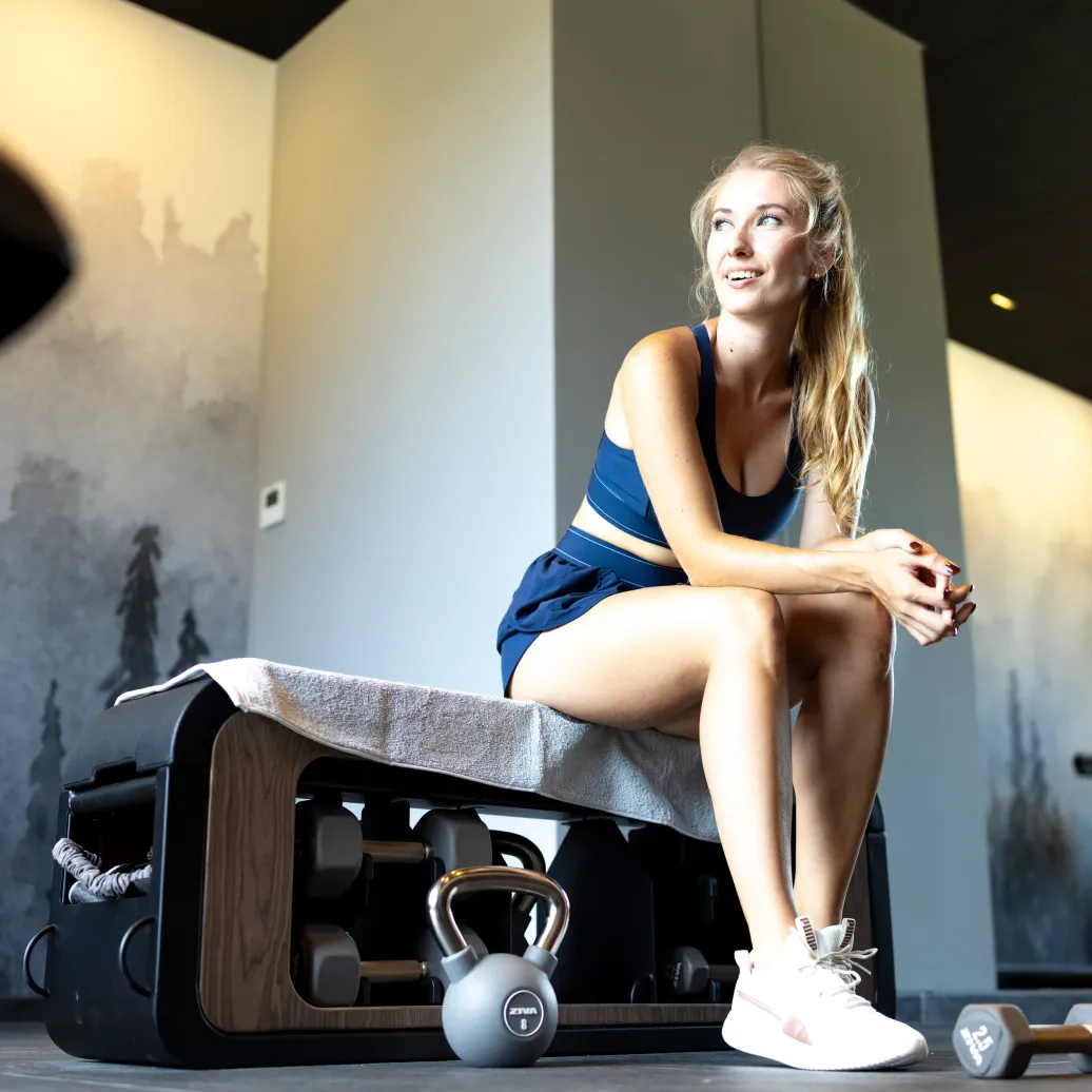 A woman sitting on a weight bench with a towel, surrounded by fitness equipment in a gym.