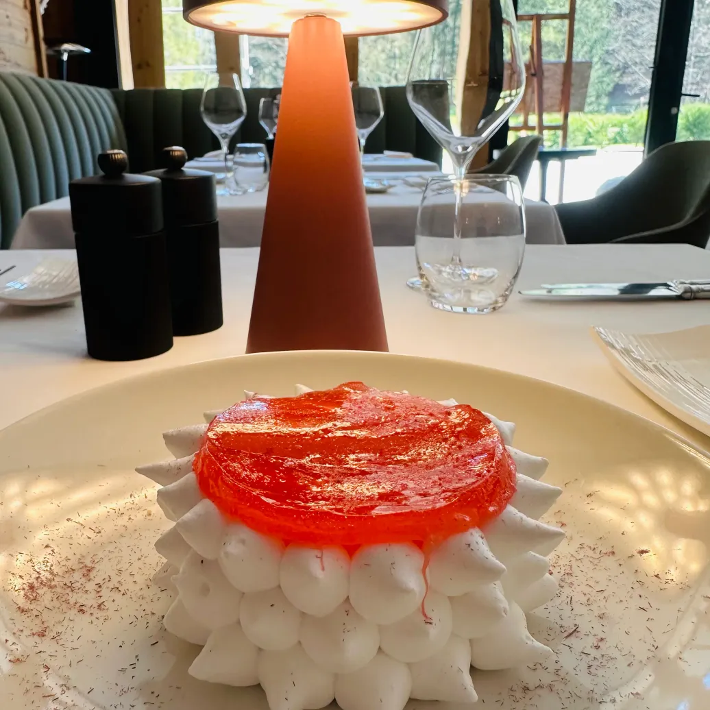 A dessert made of stacked meringues covered with red jelly, placed on a white plate with chocolate shards, in a restaurant with visible tables and chairs in the background.