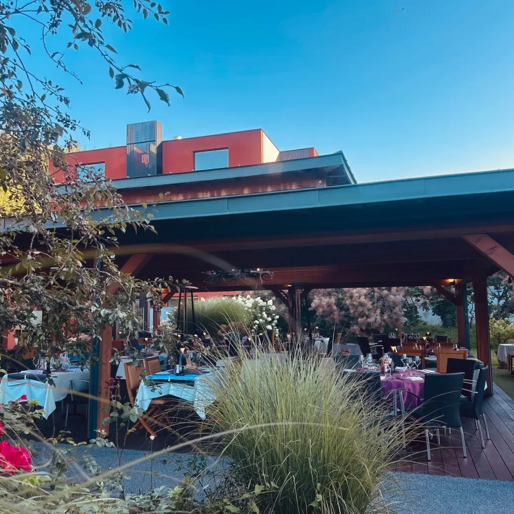 A covered terrace with tables set for a meal, surrounded by vegetation and flowers, under a clear sky.
