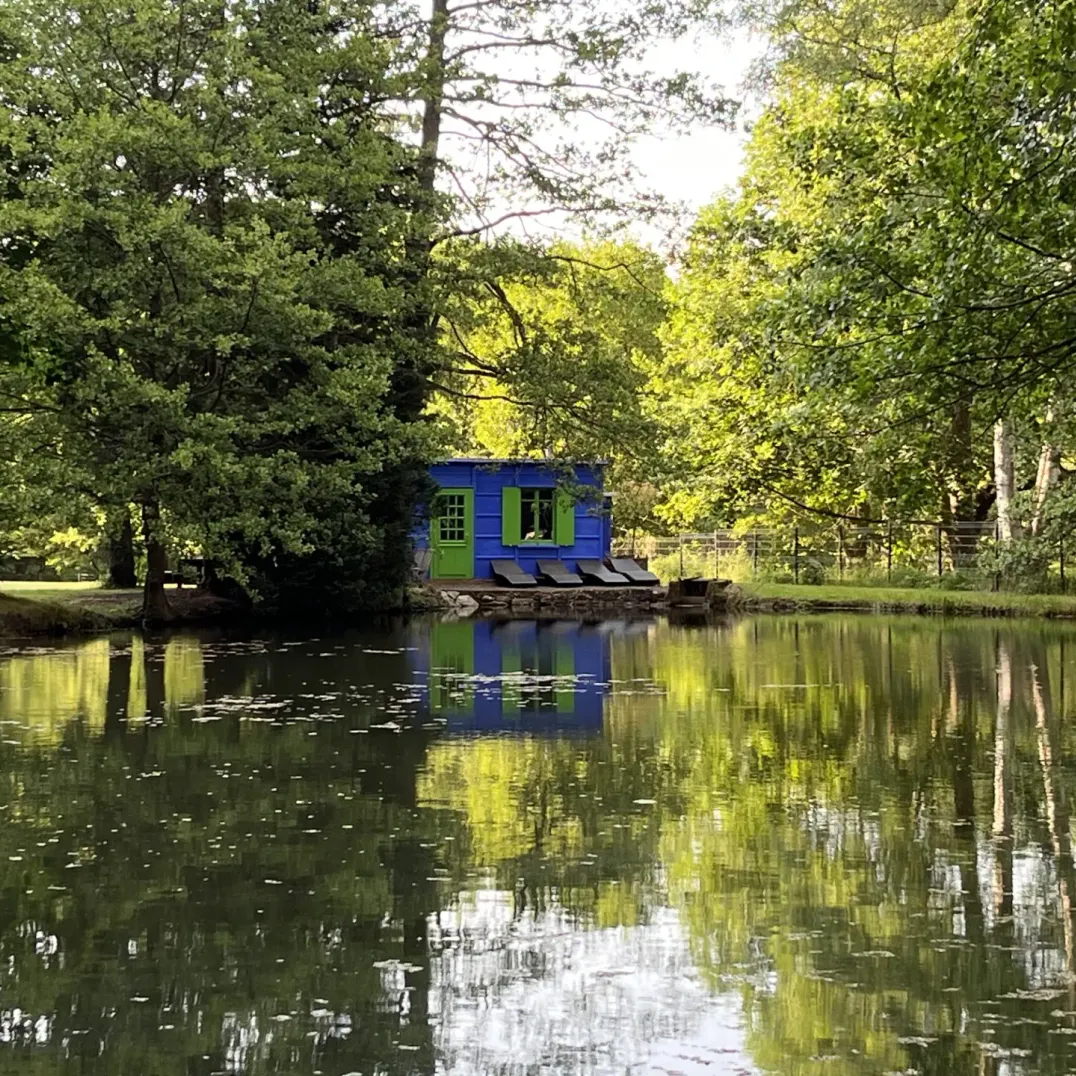 Une petite maison bleue avec des volets verts est située au bord d'un étang entouré d'arbres