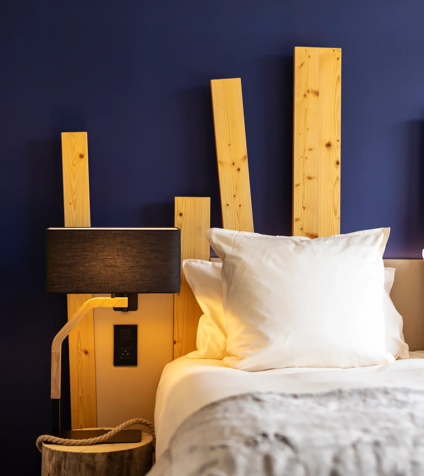 Bett mit einem hellen Holzkopfteil, zwei weißen Kissen und einer grauen Decke, einer schwarzen Nachttischlampe und einem Holzkorb daneben