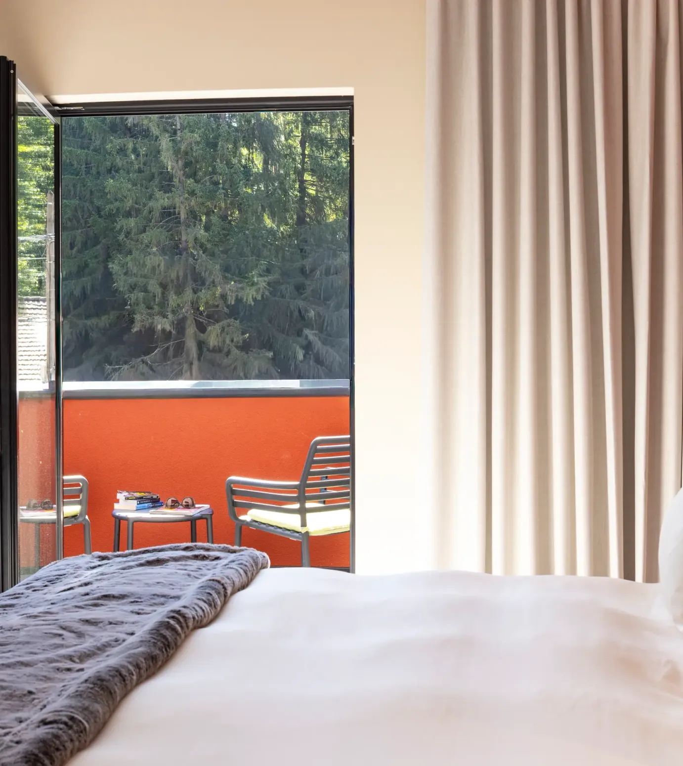 Zimmer mit einem Bett und einer offenen Glastür zum Balkon mit Stühlen und einem Tisch, Blick auf Bäume