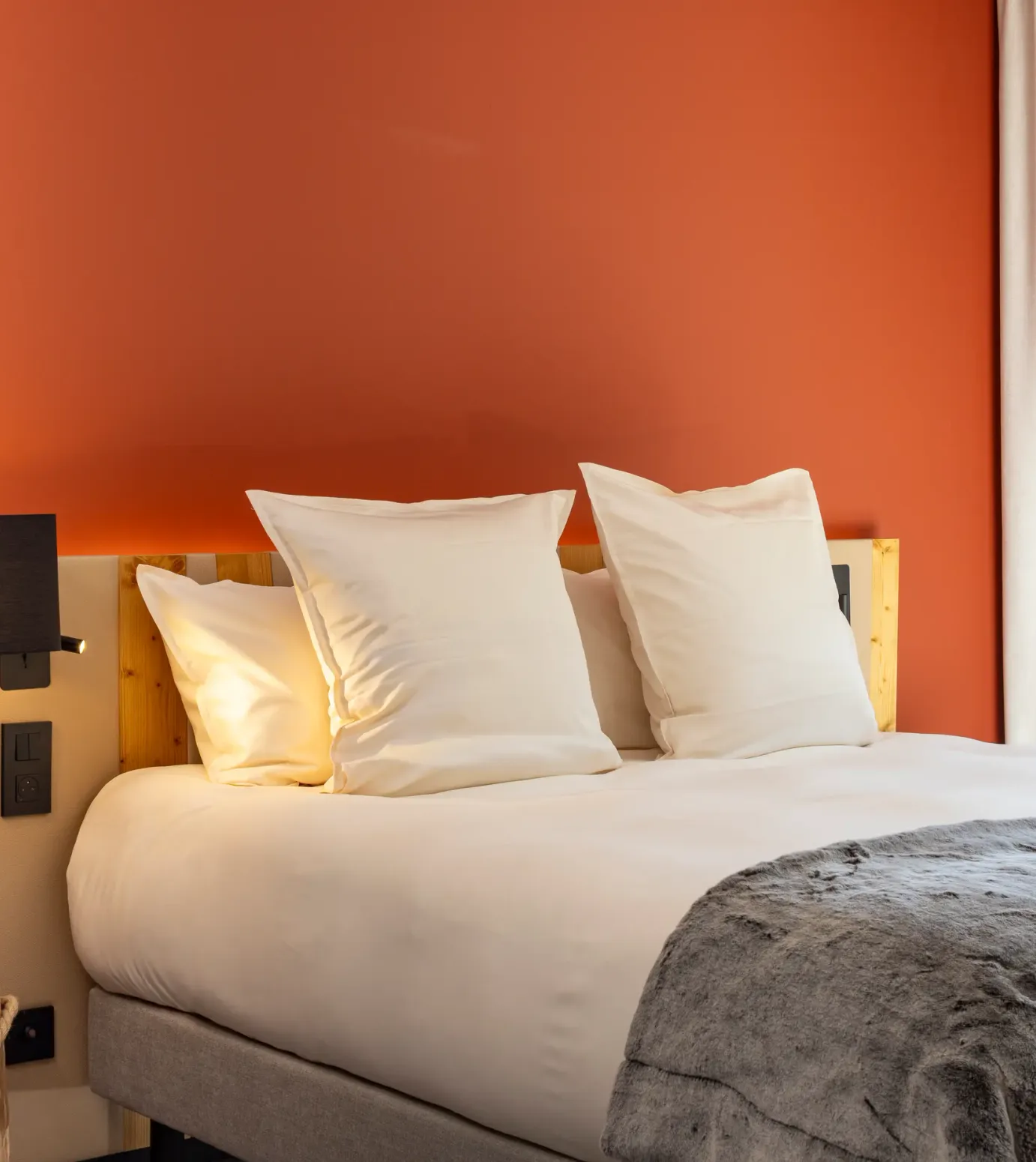 Bett mit zwei weißen Kissen, einer Holz-Wandleuchte und einer grauen Decke, vor einer orangen Wand und einem Fenster mit weißen Vorhängen