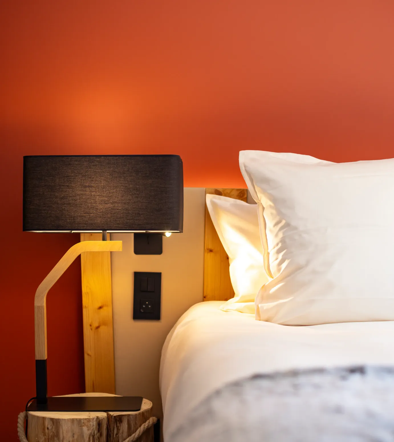 Bed with a light wood headboard, two white pillows, and a bedside lamp with a black shade on a raw wood nightstand.