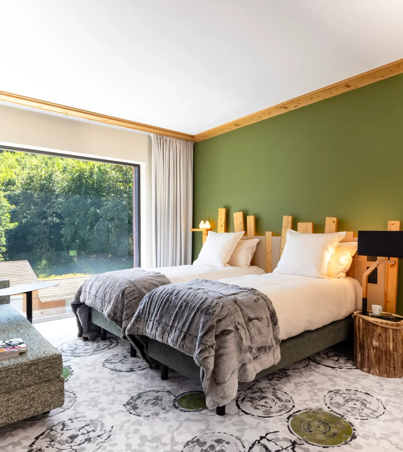 Zimmer mit zwei Betten, einer Liege, einem Holznachttisch und einem großen Fenster mit Blick auf einen Wald