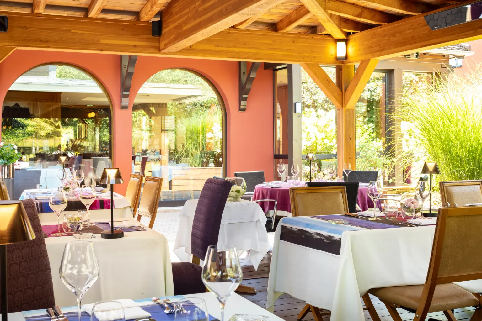Restaurantterrasse mit gedeckten Tischen, weißen Tischdecken und Weingläsern unter einer Holzkonstruktion mit orangen Bögen