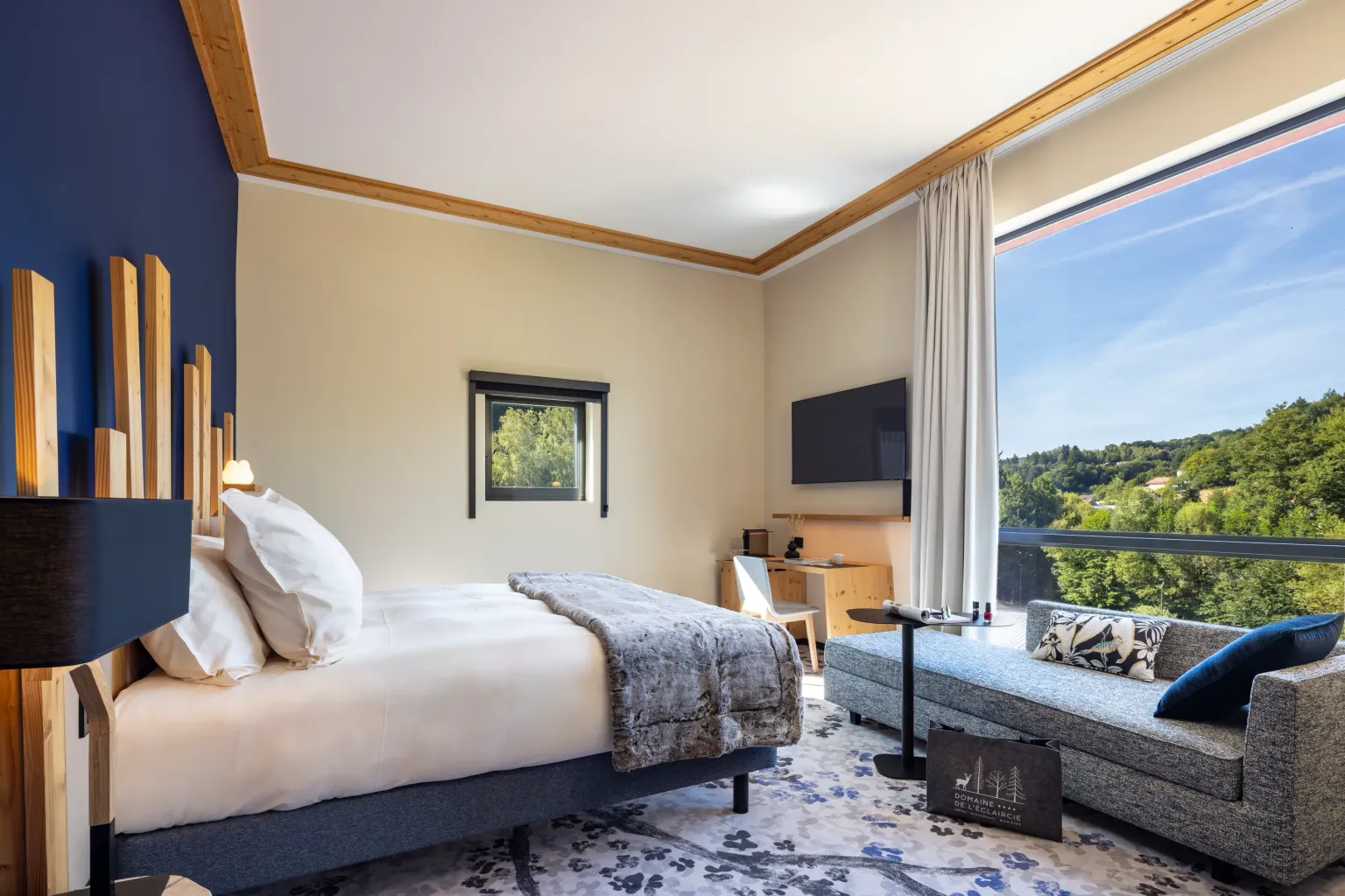 Room with a bed, a bedside table, a sofa, and a large window offering a view of a forest.