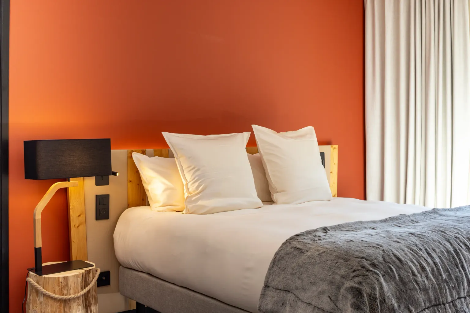 Bed with two white pillows, a wooden wall lamp, and a gray blanket, in front of an orange wall and a window with white curtains.