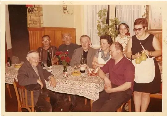 Un groupe de personnes est assis autour d'une table à manger, avec des bouteilles, des verres et des fleurs, dans une pièce avec des rideaux et une cheminée.