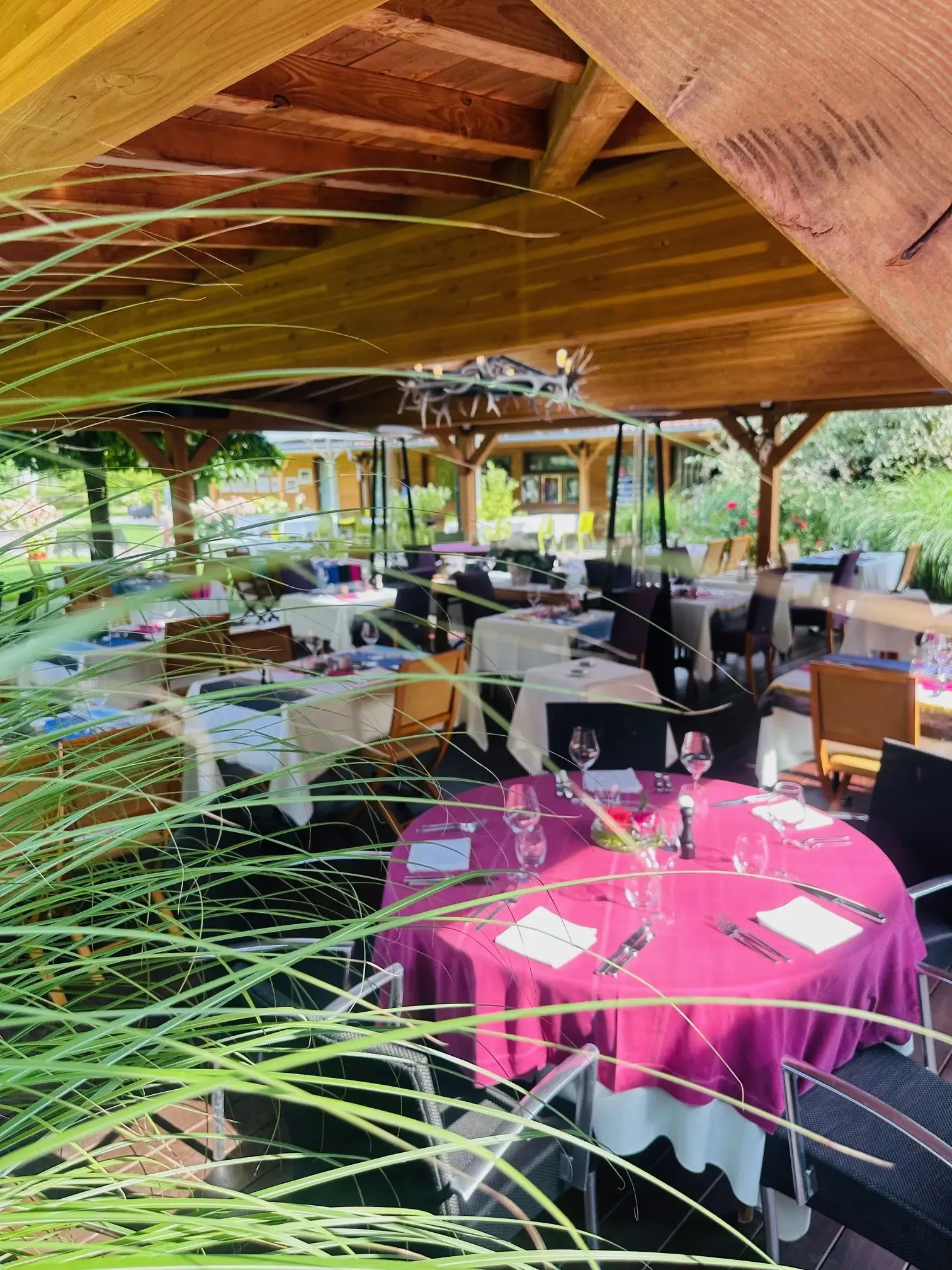 Une terrasse couverte avec des tables dressées de nappes roses et de verres à vin, entourée de végétation