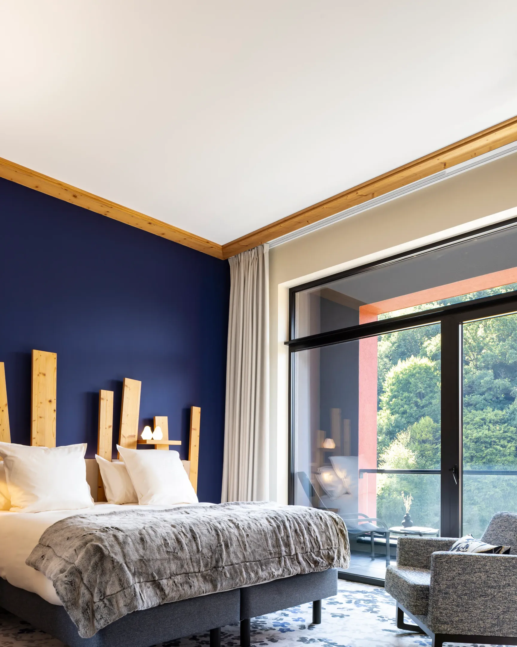 Chambre avec un lit double recouvert d'une couverture grise, une tête de lit en bois, une grande fenêtre donnant sur une forêt et un fauteuil à côté