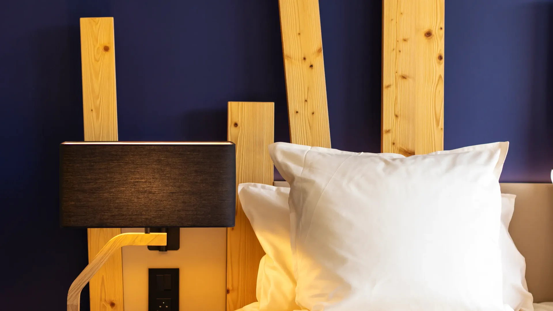 Bett mit einem hellen Holzkopfteil, zwei weißen Kissen und einer grauen Decke, einer schwarzen Nachttischlampe und einem Holzkorb daneben