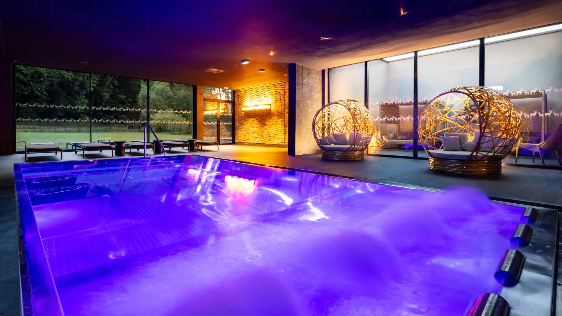 Piscine intérieure éclairée en violet avec des chaises longues et des fauteuils sphériques en osier illuminés à proximité, entourée de grandes baies vitrées donnant sur un jardin