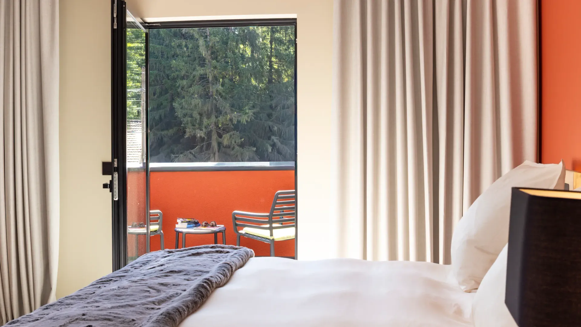 Room with a bed and an open glass door leading to a balcony with chairs and a table, view of trees