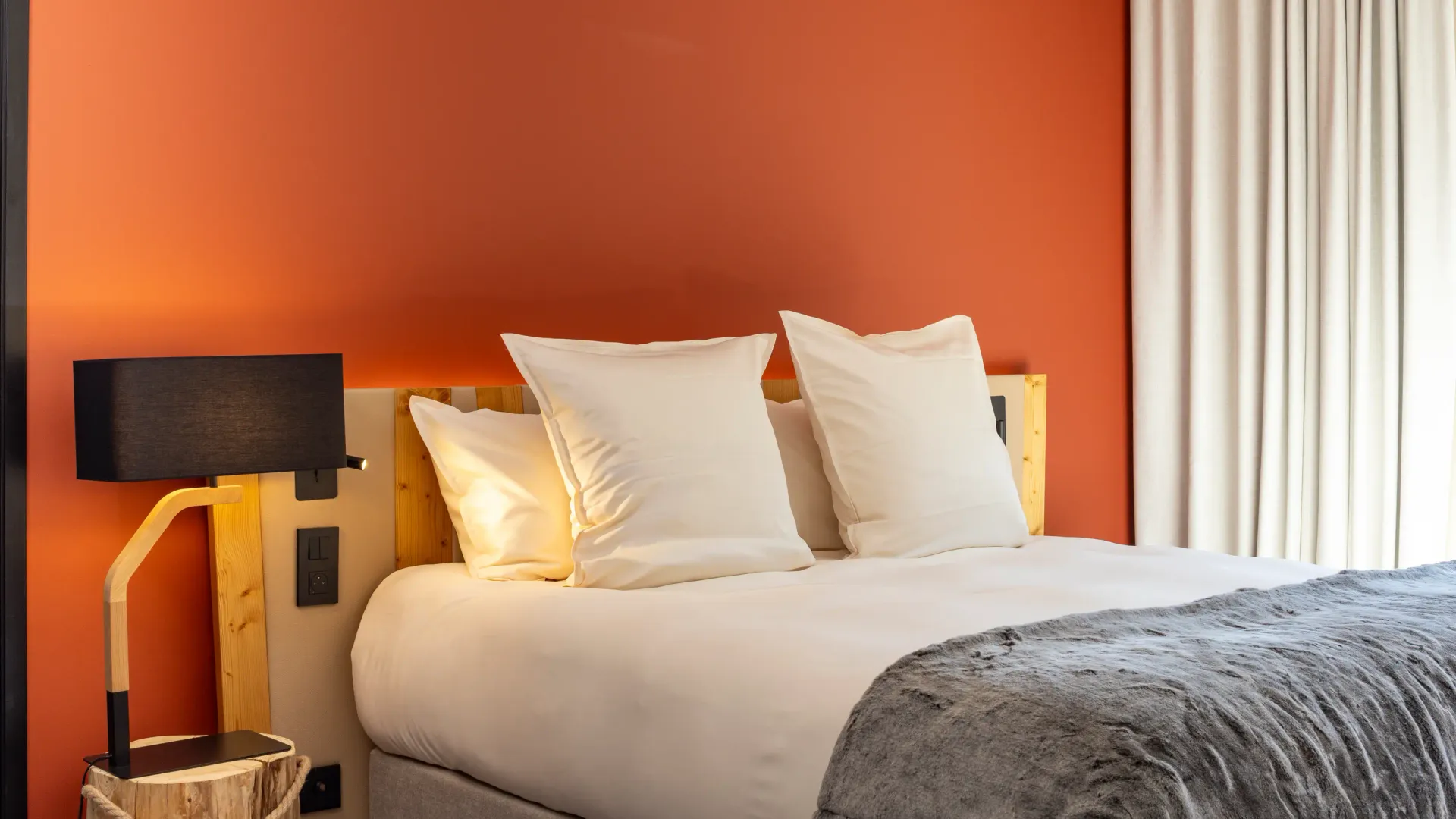 Lit avec deux oreillers blancs, une lampe murale en bois et une couverture grise, devant un mur orange et une fenêtre avec des rideaux blancs