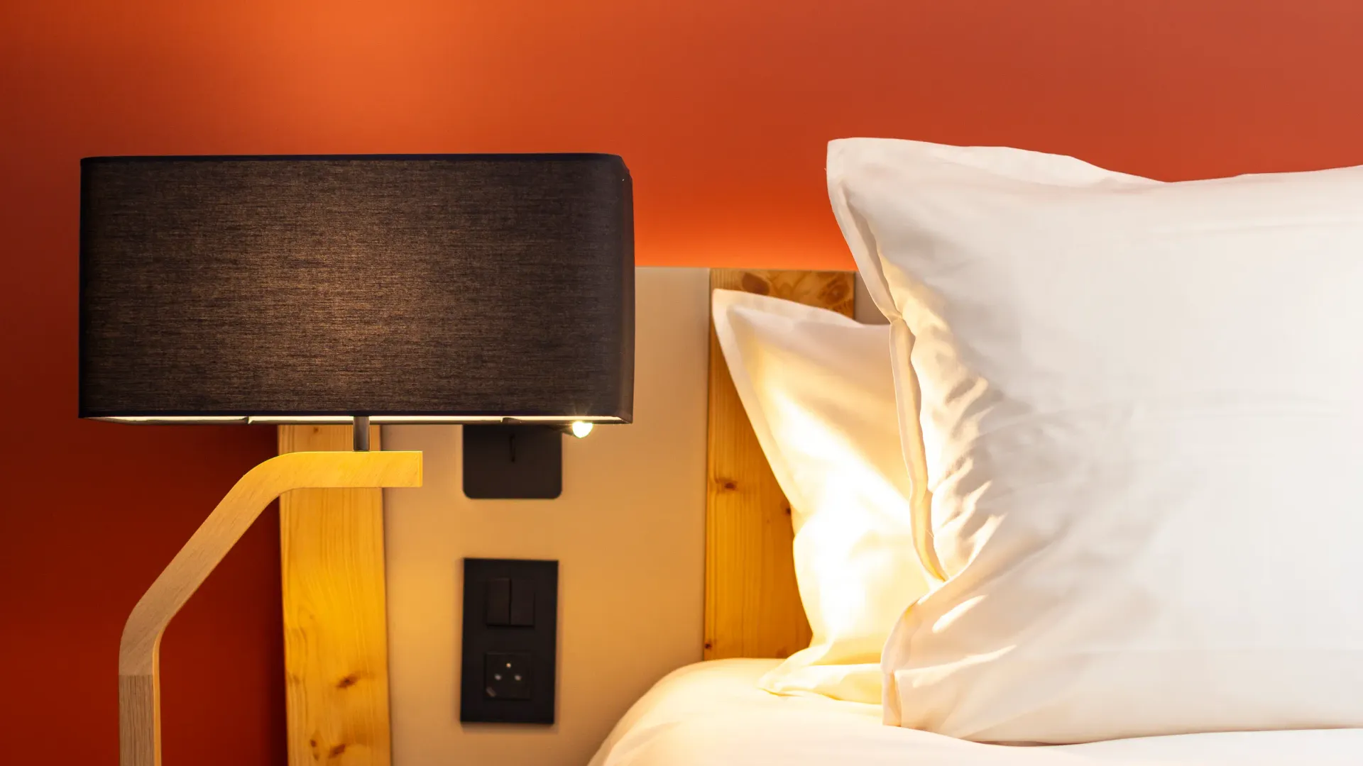 Lit avec une tête de lit en bois clair, deux oreillers blancs et une lampe de chevet à abat-jour noir sur une table de nuit en bois brut