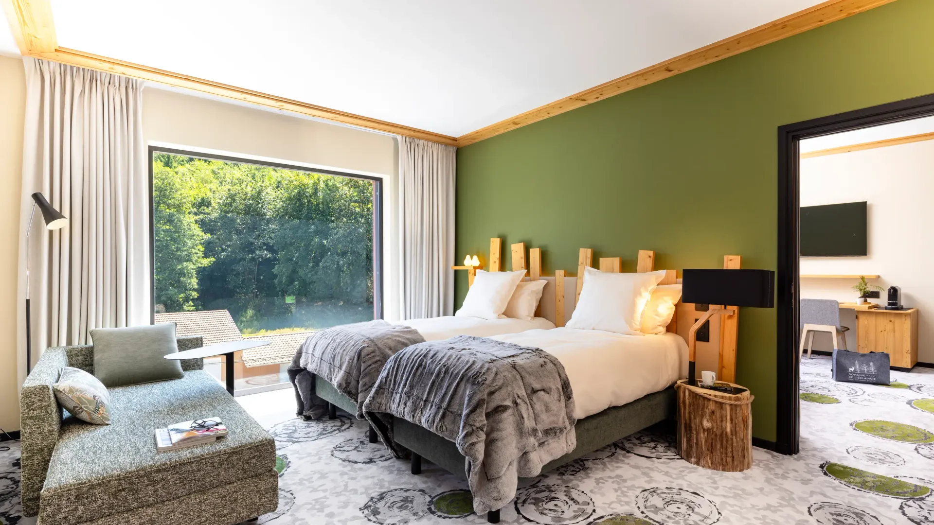 Zimmer mit zwei Betten, einer Liege, einem Holznachttisch und einem großen Fenster mit Blick auf einen Wald