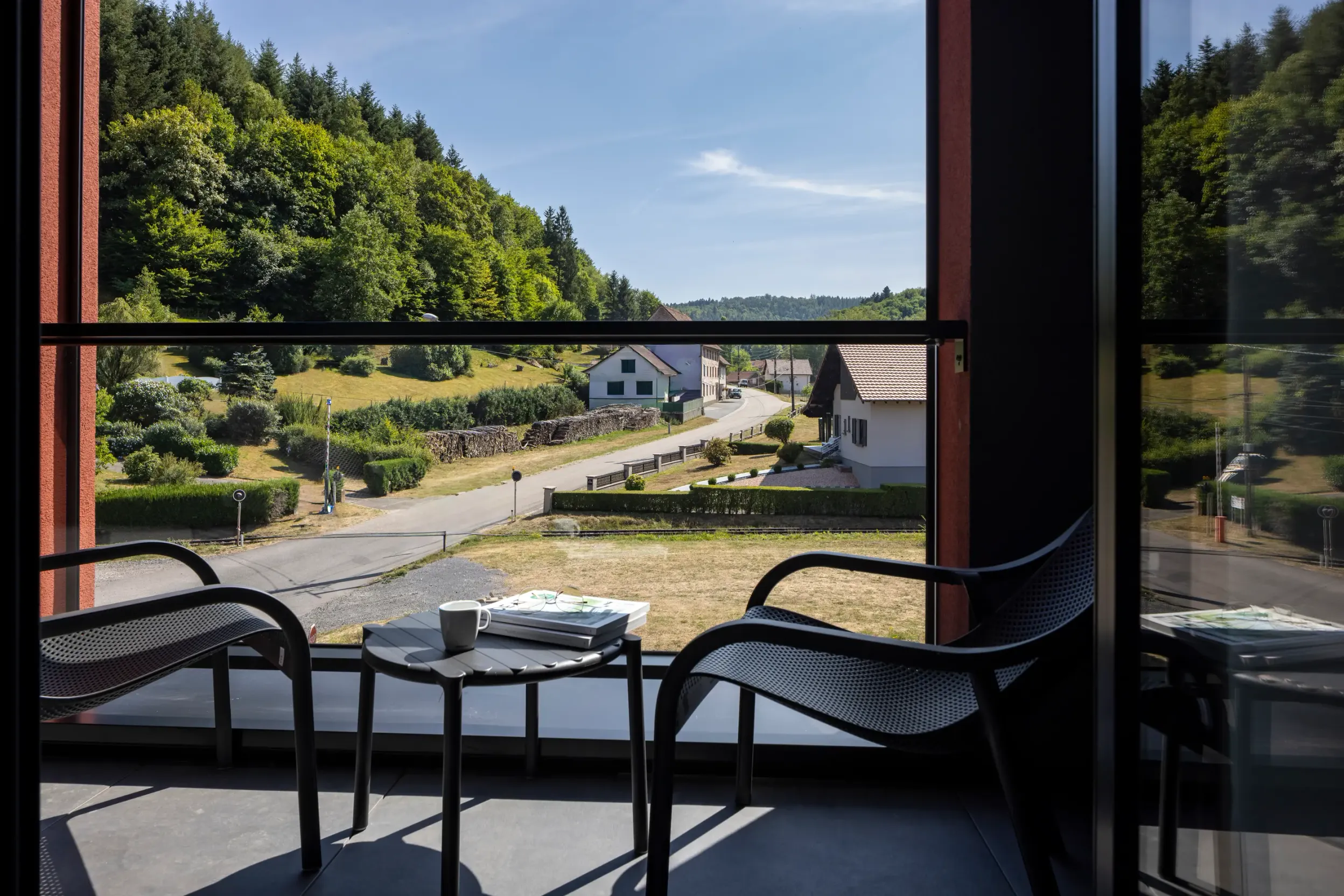 Aus einem offenen Fenster auf einen Balkon mit Stühlen und einem Tisch, der eine Landstraße mit Häusern und Grünflächen unter einem klaren Himmel zeigt