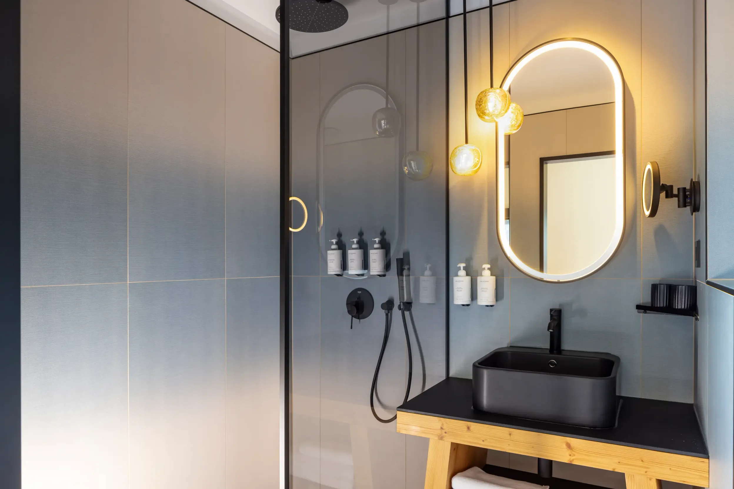 Moderne Badezimmer mit ovalem Spiegel, schwarzem Waschbecken auf Holzschrank und aufgereihten Toilettenartikeln