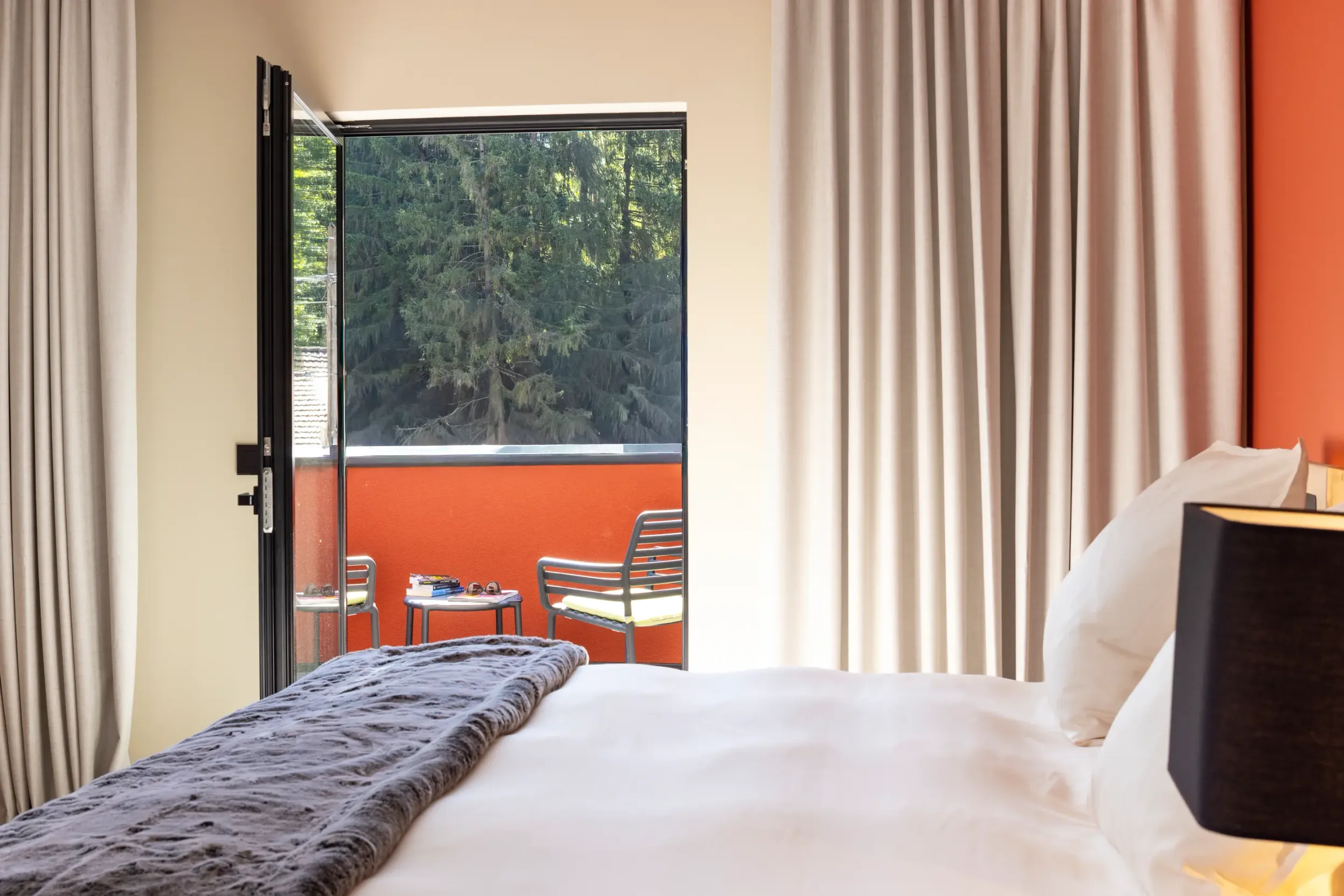 Chambre avec un lit et une porte vitrée ouverte donnant sur un balcon avec des chaises et une table, vue sur des arbres