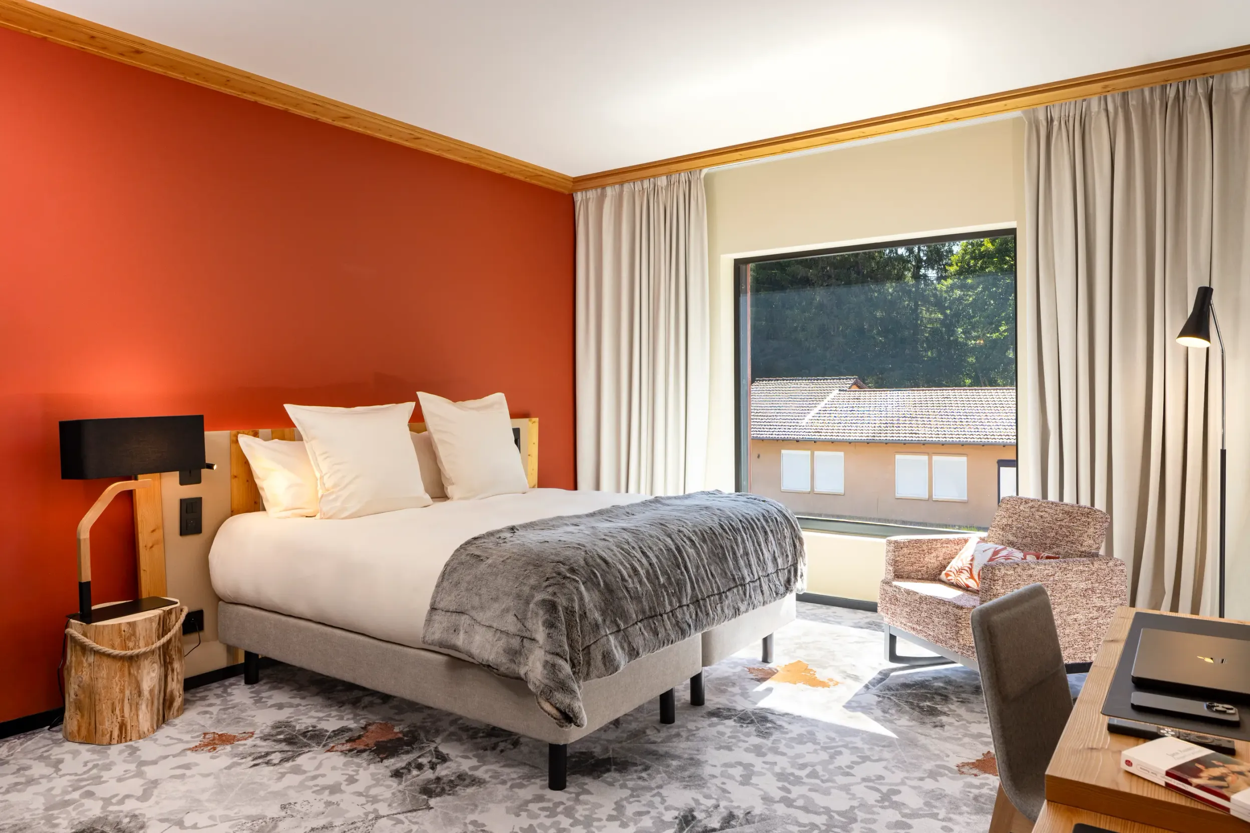 Chambre avec un lit double, une table de chevet, une lampe, un fauteuil, un bureau et une grande fenêtre donnant sur un bâtiment extérieur