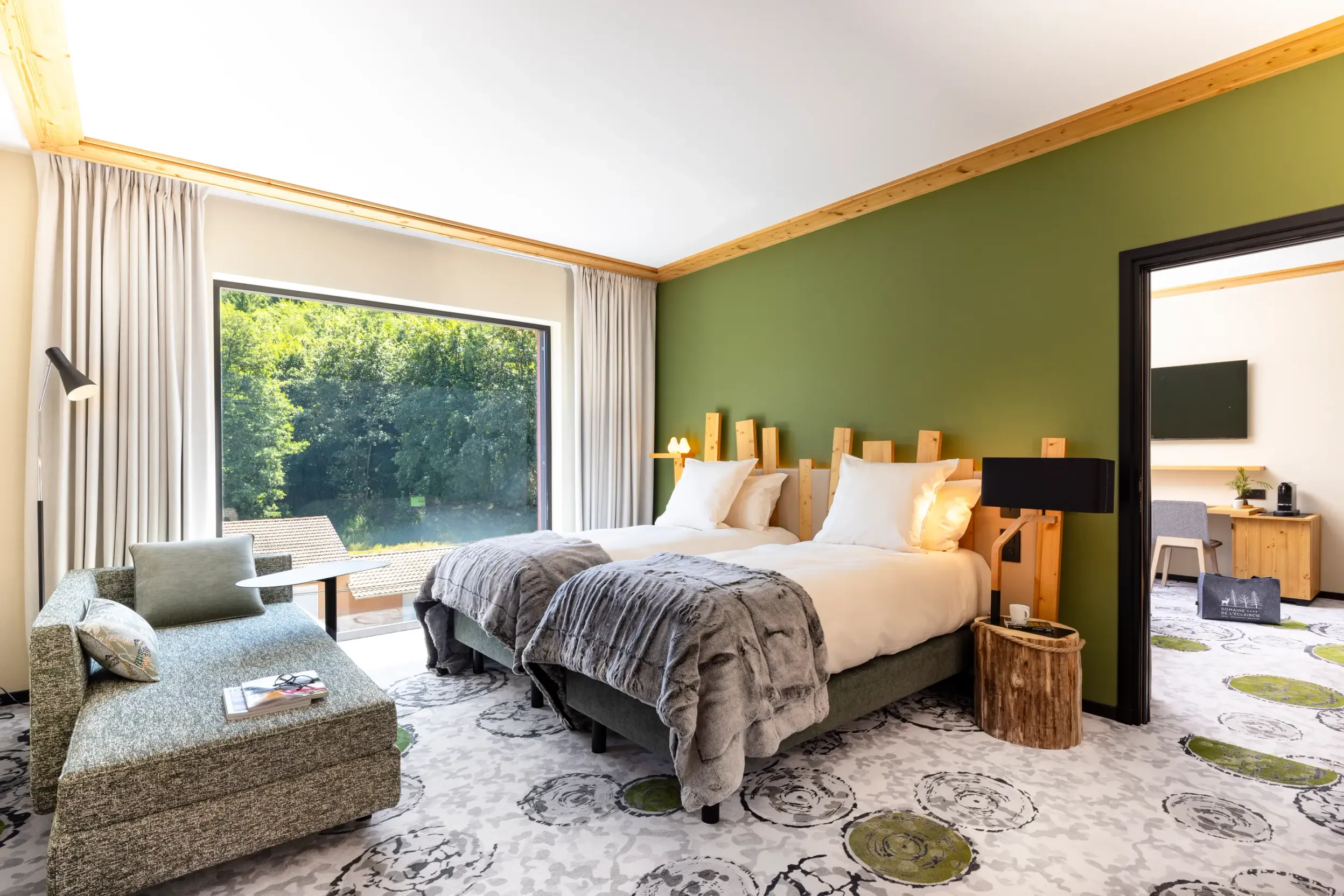 Zimmer mit zwei Betten, einer Liege, einem Holznachttisch und einem großen Fenster mit Blick auf einen Wald