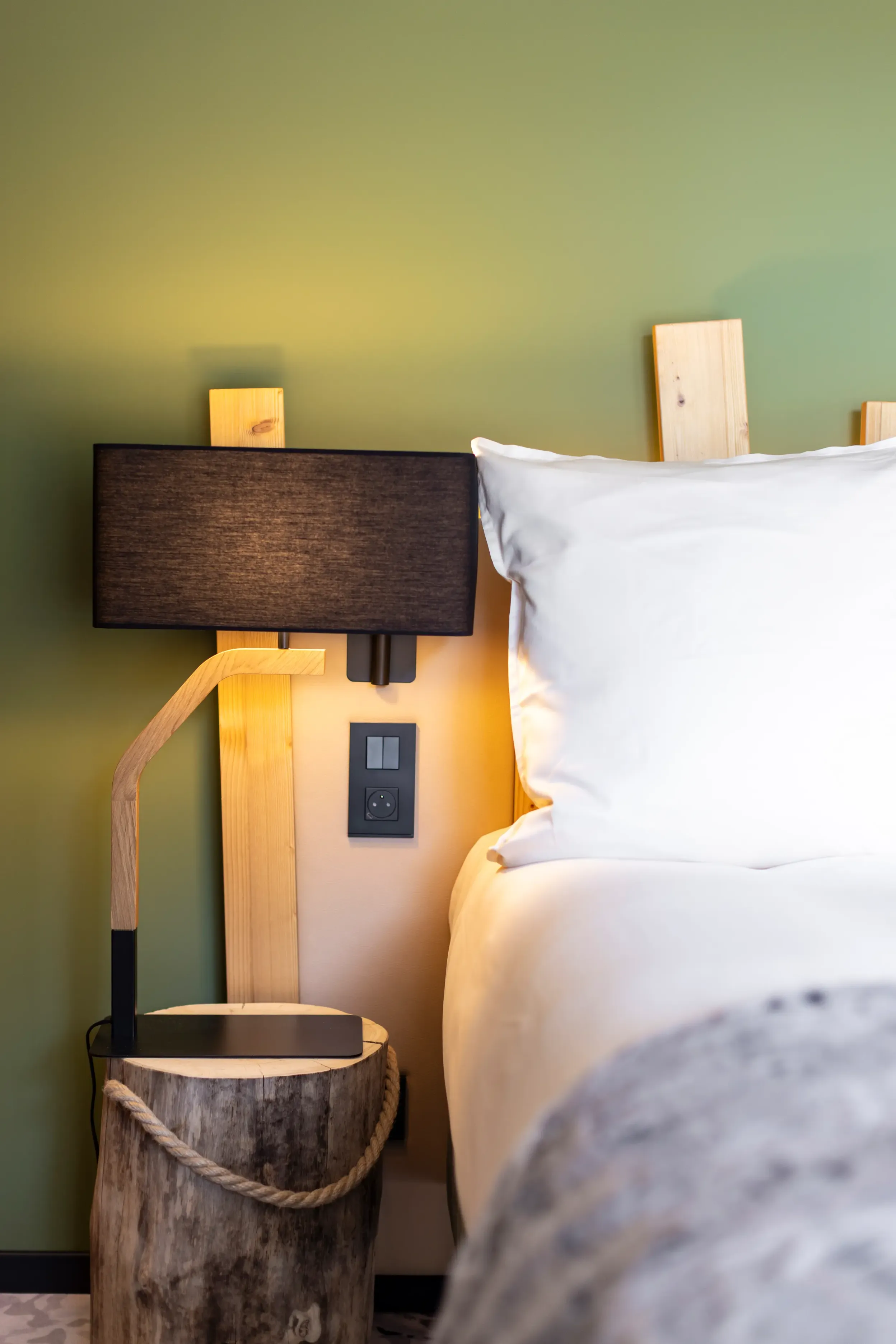 Eine Stehlampe aus Holz mit einem schwarzen Lampenschirm steht neben einem Bett mit einem hellen Holzkopfteil und weißen Kissen.