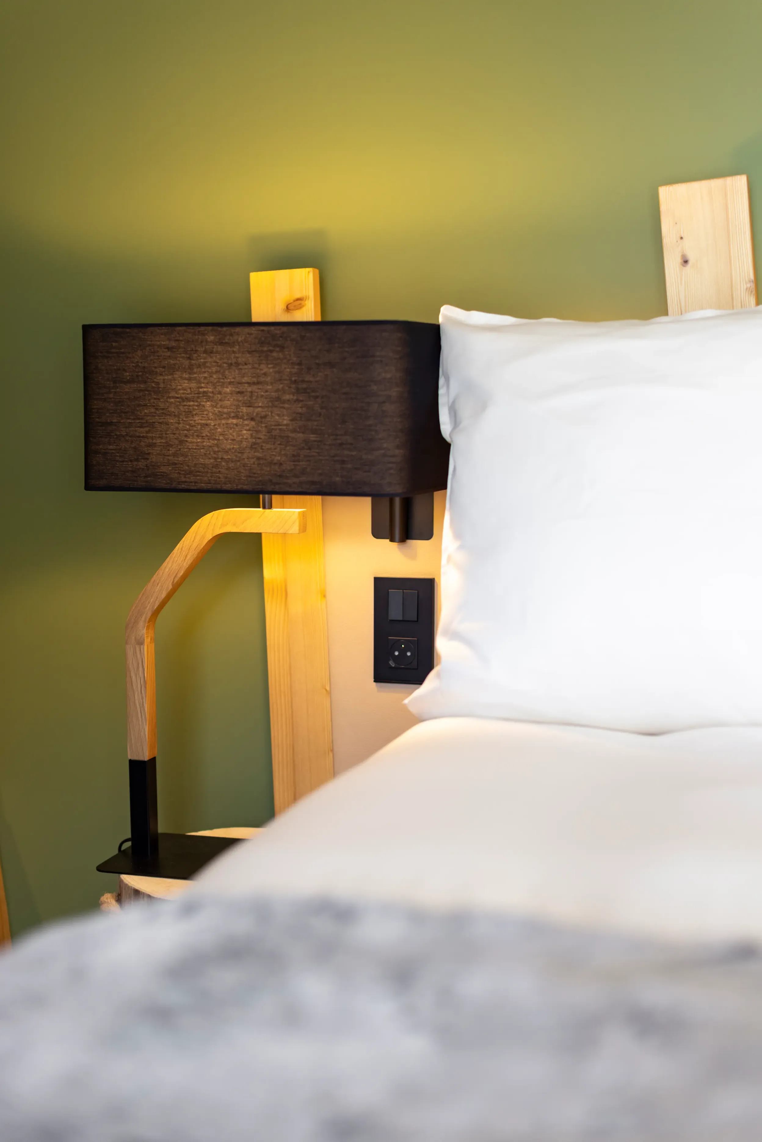 Wooden bedside lamp with black shade next to a bed with a white blanket and a pillow