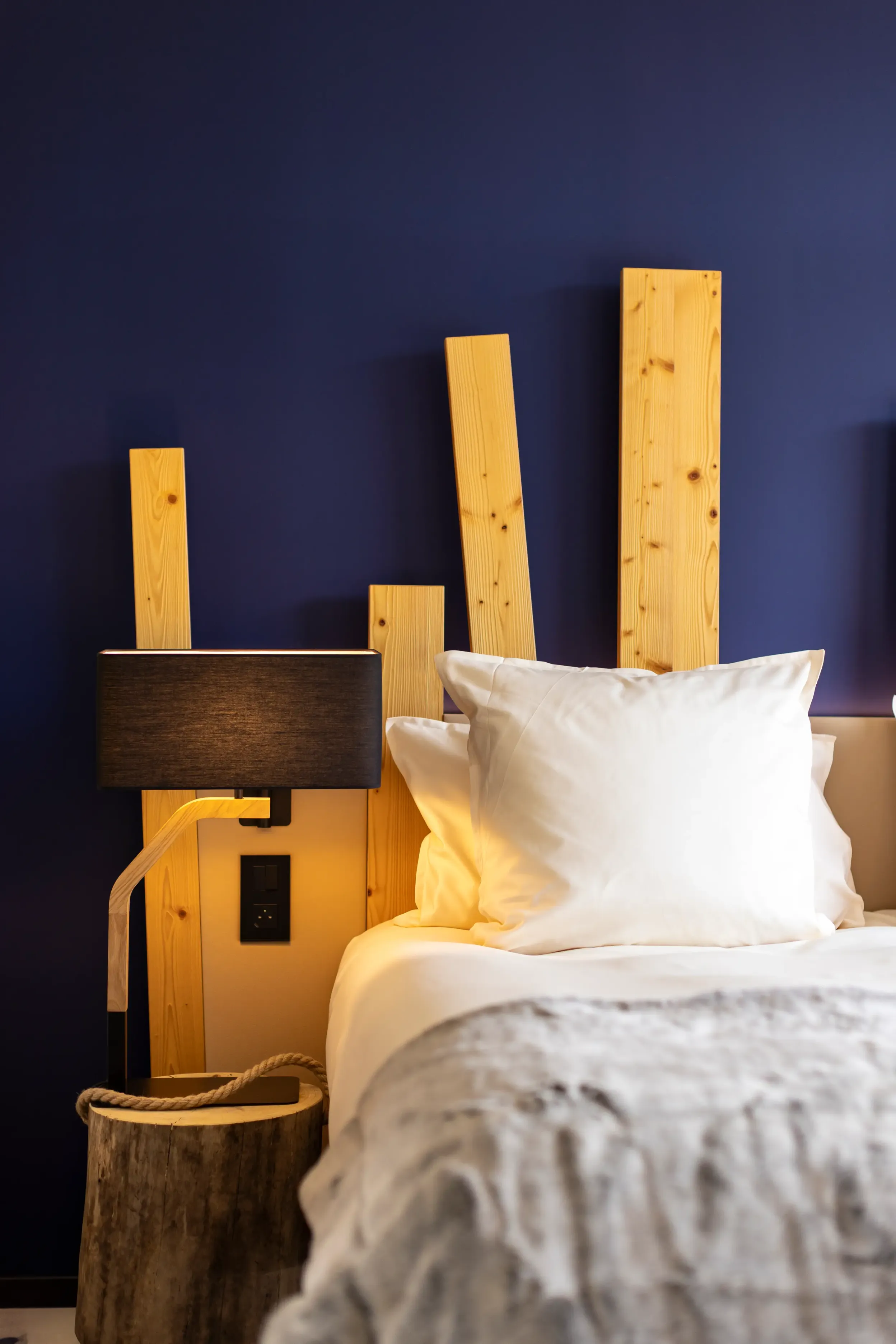 Bett mit einem hellen Holzkopfteil, zwei weißen Kissen und einer grauen Decke, einer schwarzen Nachttischlampe und einem Holzkorb daneben